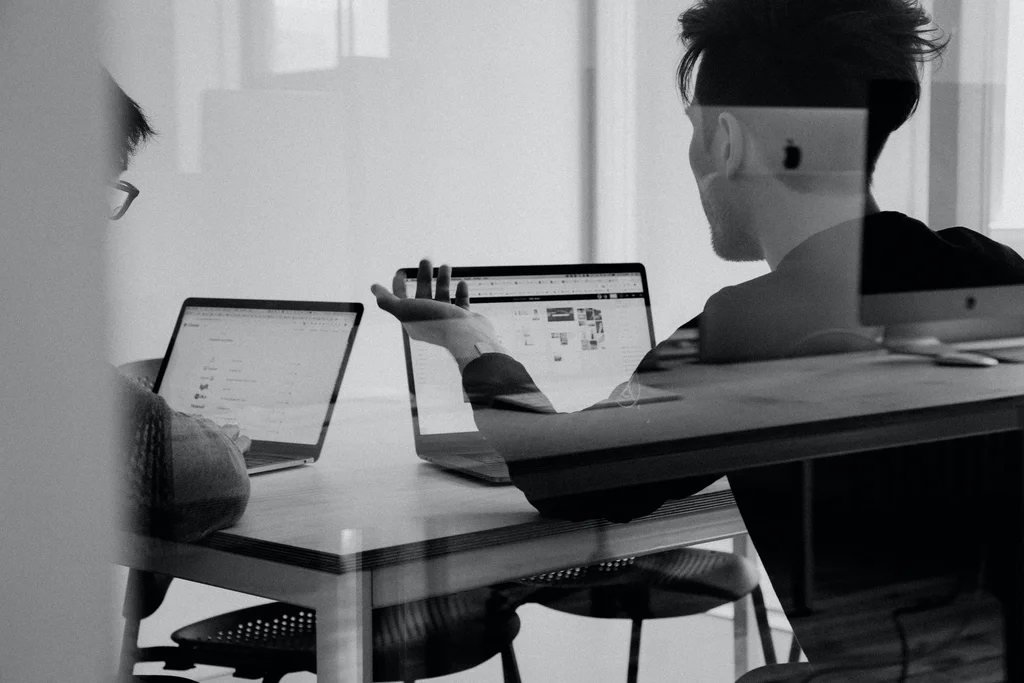 An image of 2 people talking while facing their laptops